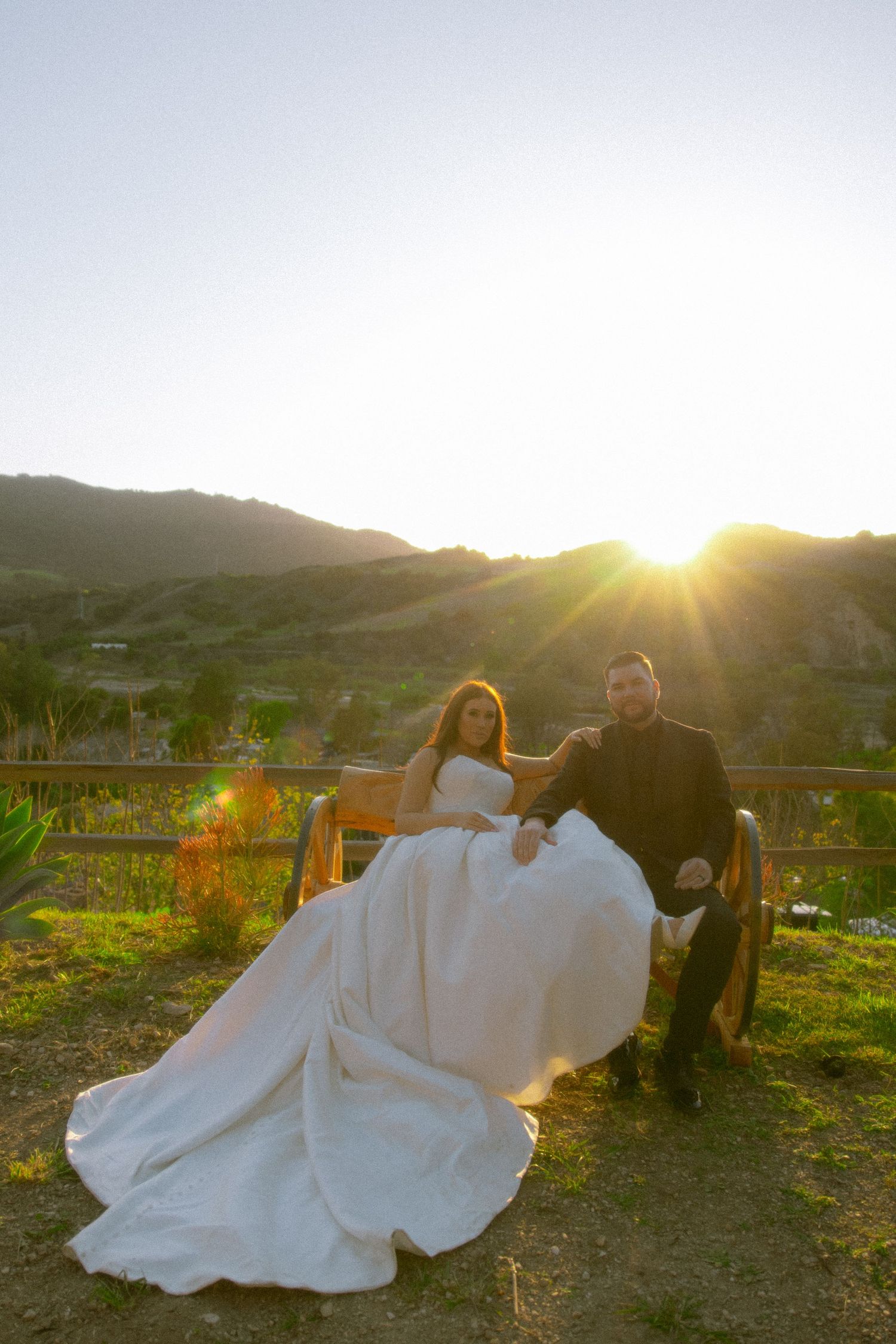 A couple in wedding attire sits on a bench at sunset with rolling hills and golden light in the background.