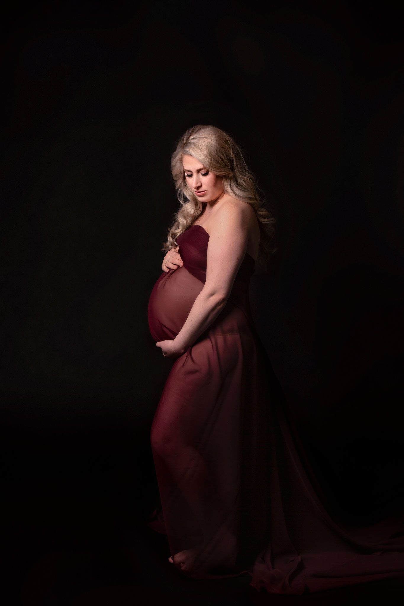 A pregnant woman in a flowing burgundy dress poses elegantly against a dark background in a maternity photo.