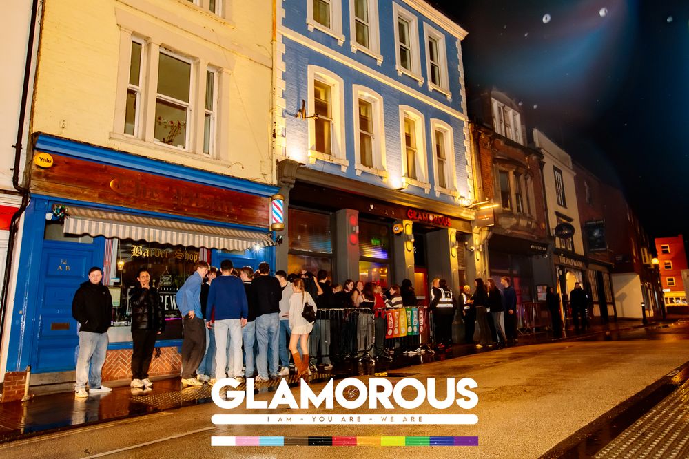 Nighttime view of people queuing outside colorful storefronts on a city street.