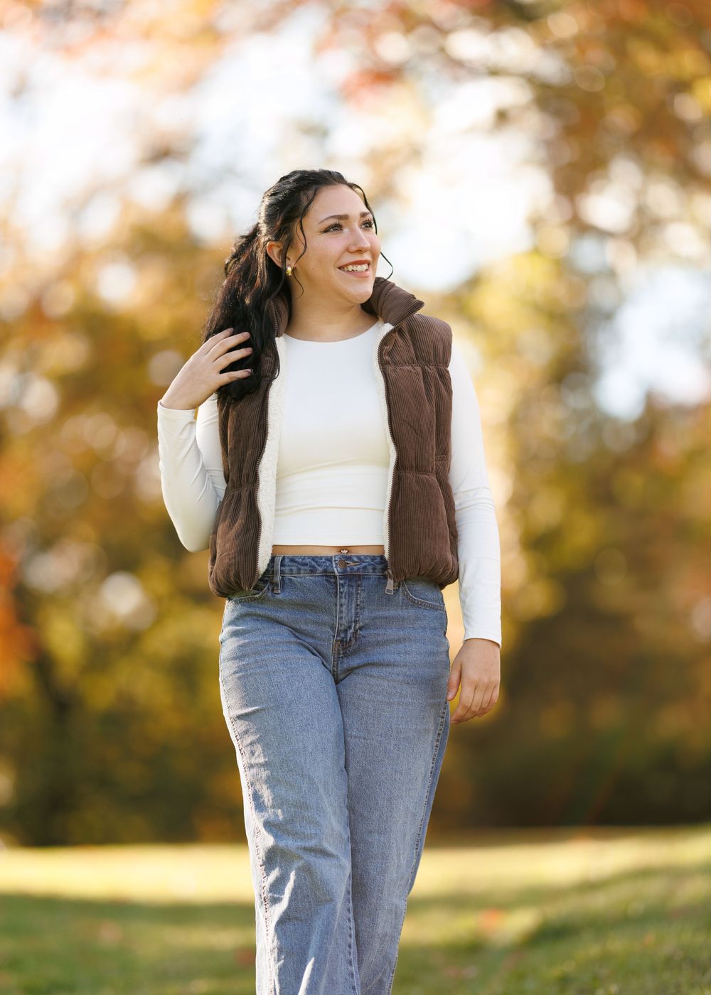 A young woman in white top and brown vest poses against autumn trees in natural lighting.