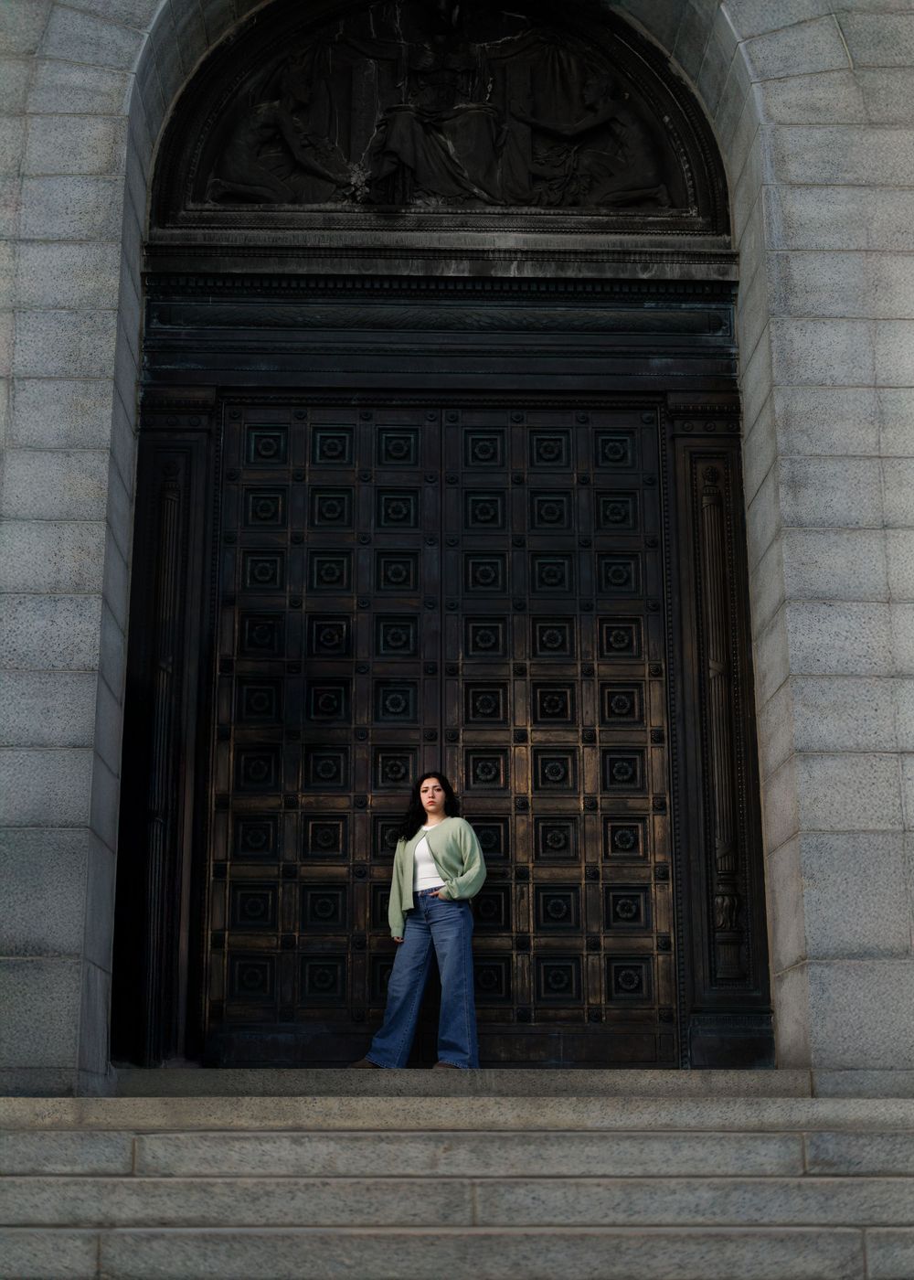 A graduate stands in an ornate stone doorway with a decorative carved wooden door and archway.