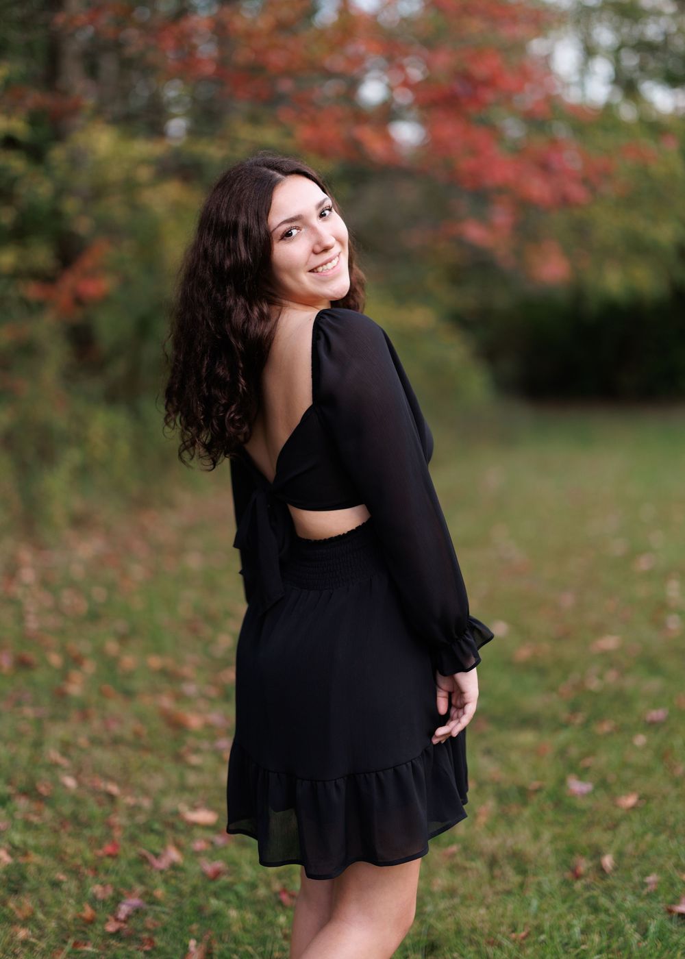 Person in black dress with cutout detail poses in autumn garden setting.