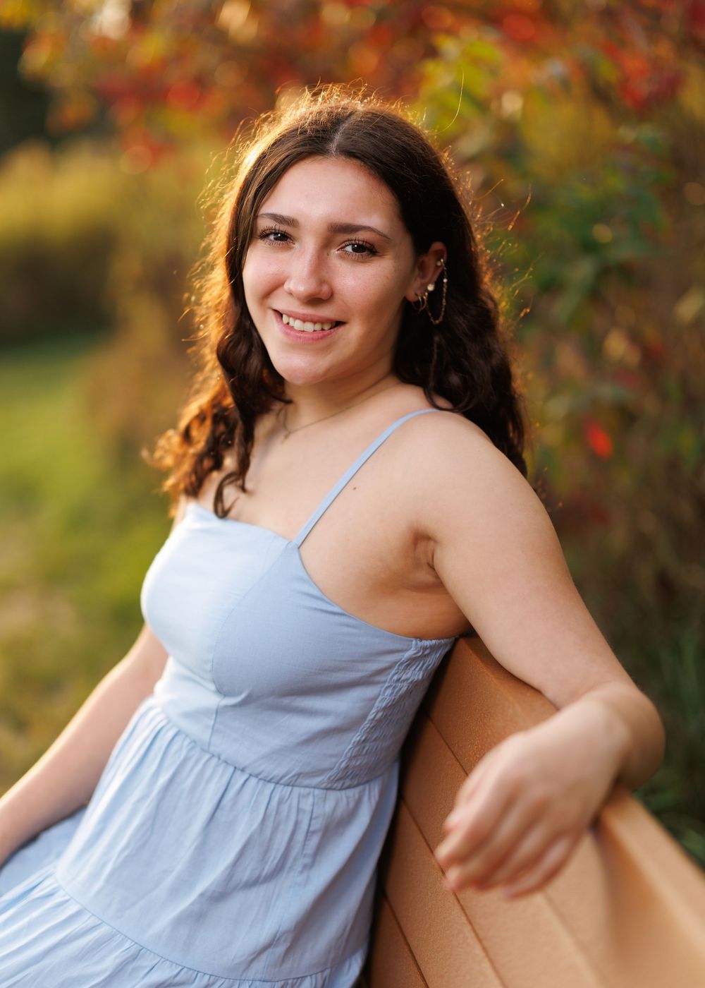 Young lady in light blue sundress poses outdoors against autumn foliage.