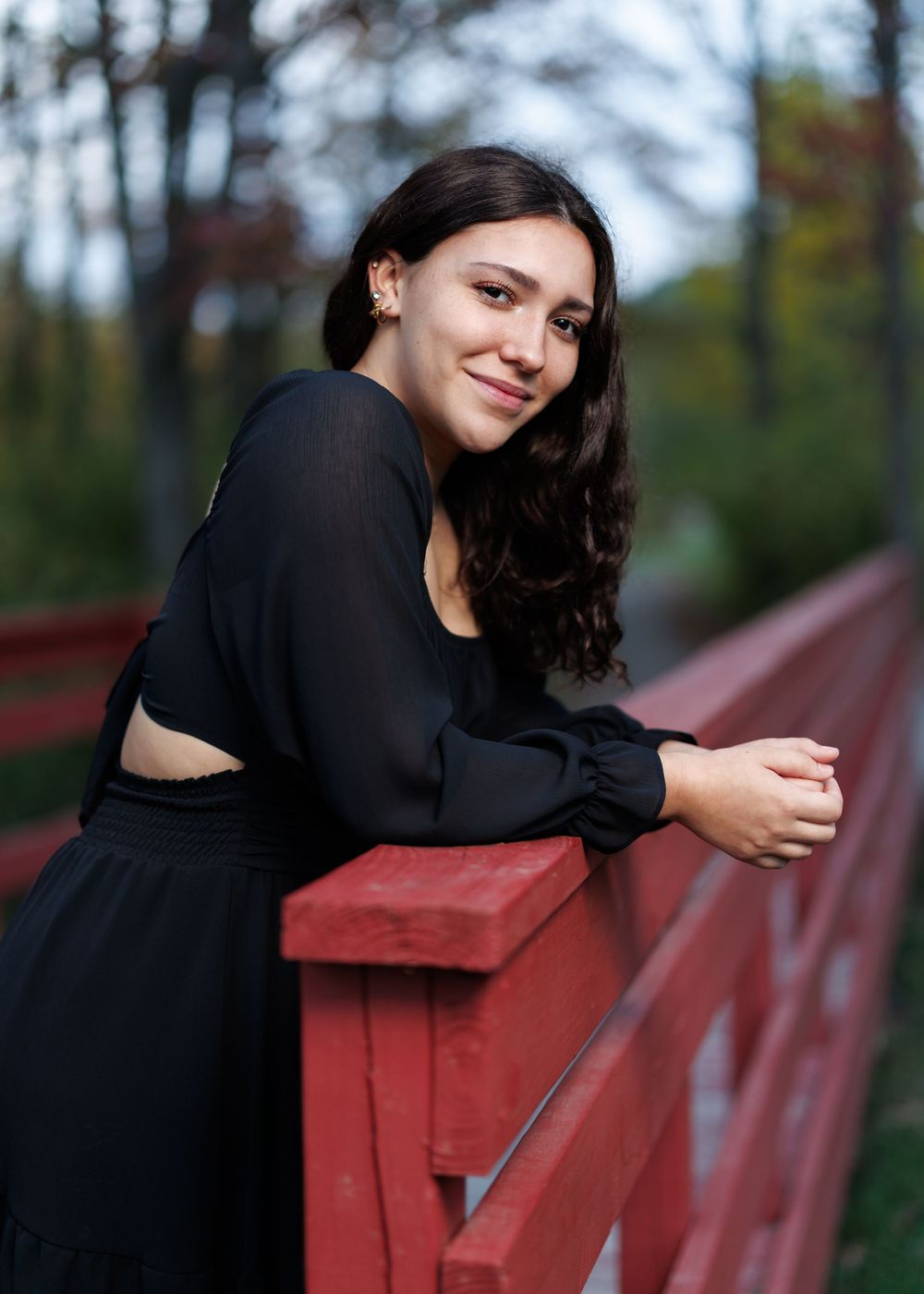 A senior in black outfit leaning on red railing during outdoor photoshoot.