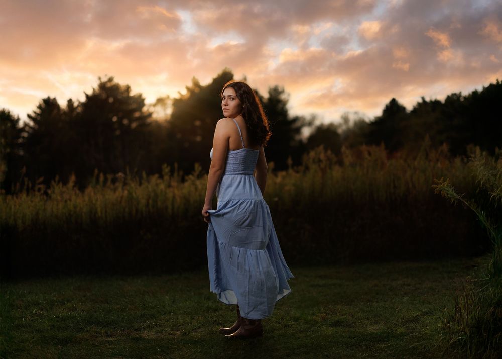 A girl in a flowing blue dress stands in a field at sunset with dramatic orange clouds in the background.