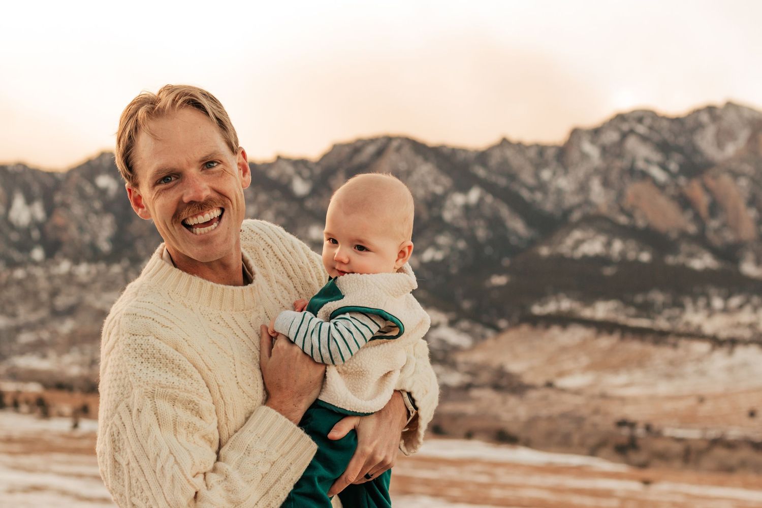 Smiling man in cream sweater holding baby outdoors mountain backdrop in winter setting | Best Family Photo Location with Mountain Views