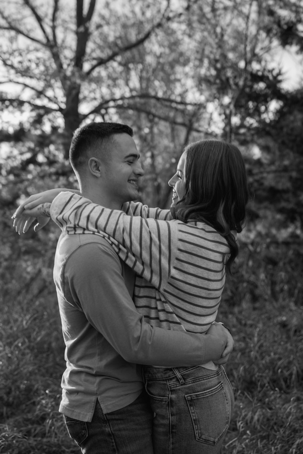 A couple in a romantic embrace outdoors, wearing casual clothing against a natural wooded background.