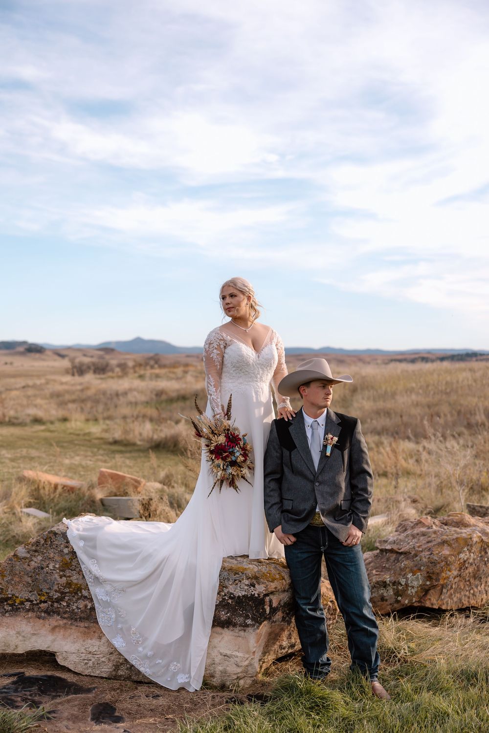 A couple in wedding attire sits together on a grassy hillside overlooking a scenic mountain landscape.