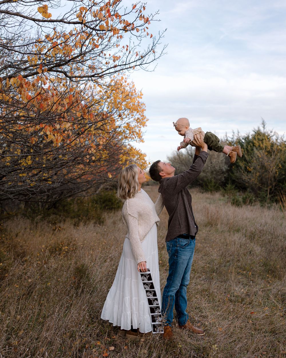 A couple dances together in a field during autumn with orange leaves on trees in the background.