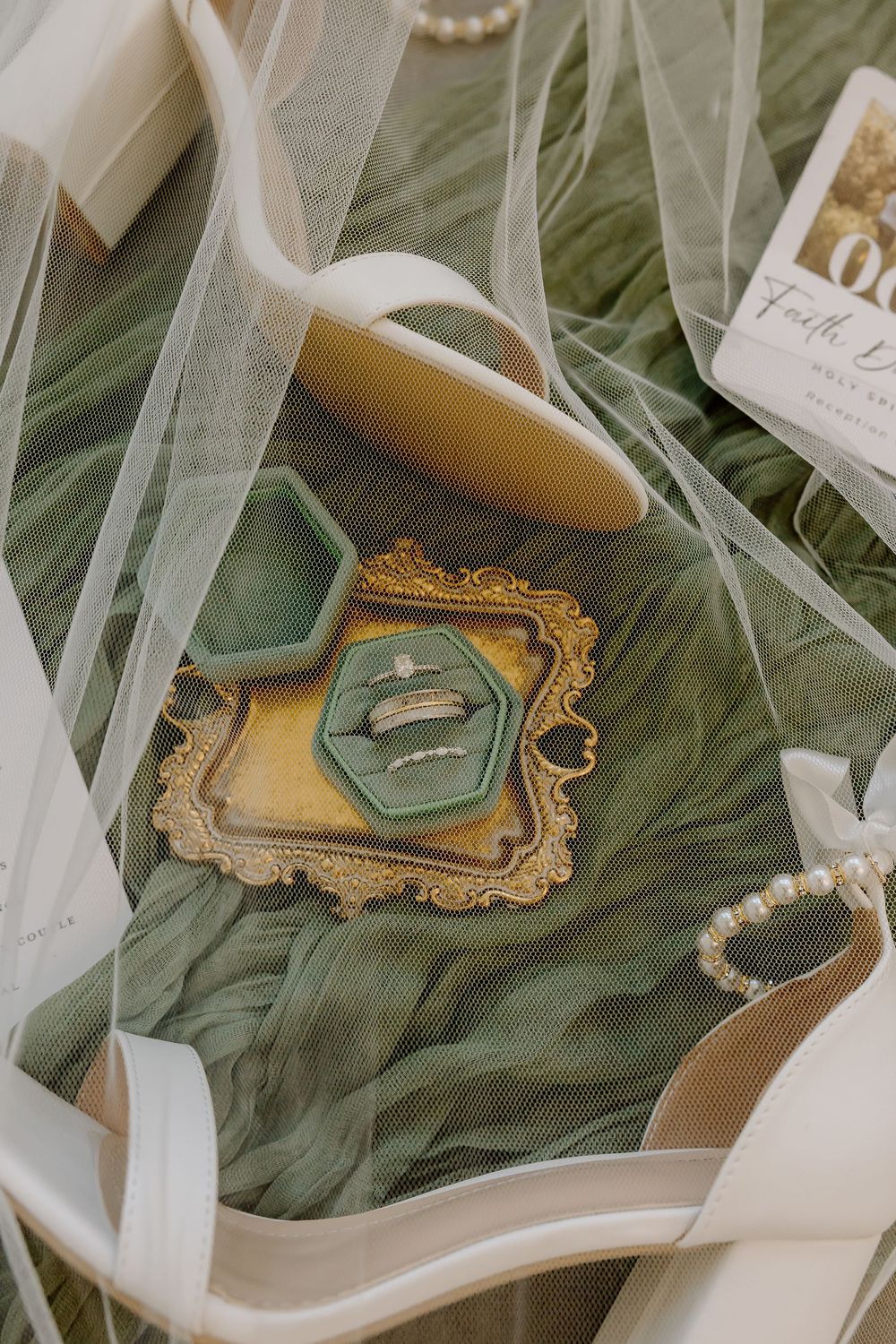 Elegant wedding rings displayed on sage green tulle fabric with ornate gold frames and white ribbons.