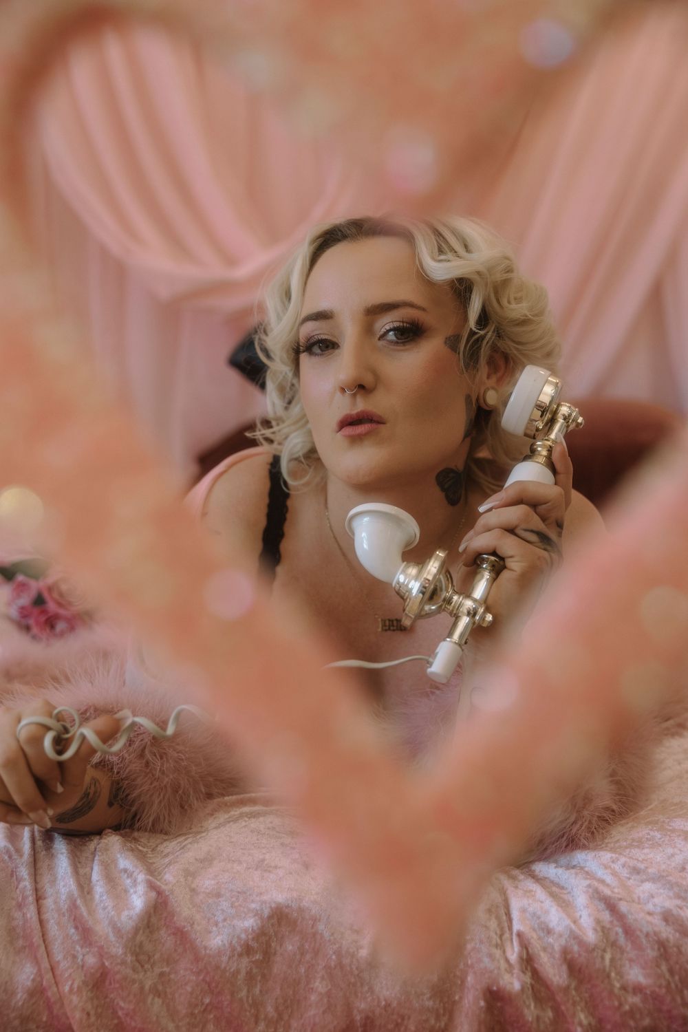 Blonde woman in pink feather robe holding vintage white and gold telephone, surrounded by pink drapery.