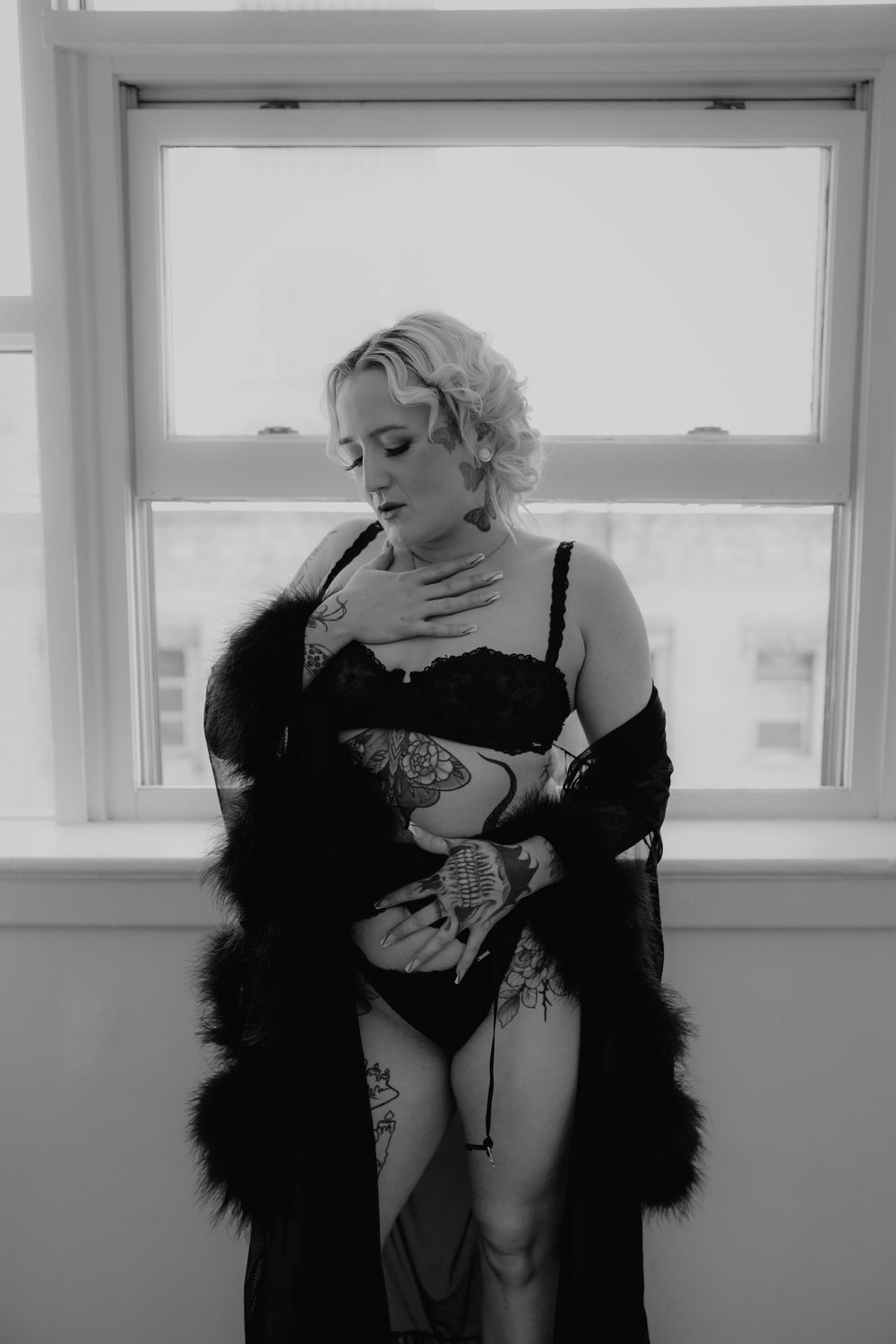 Blonde tattooed woman in black lace lingerie and fur robe poses by a window in black and white boudoir photo.