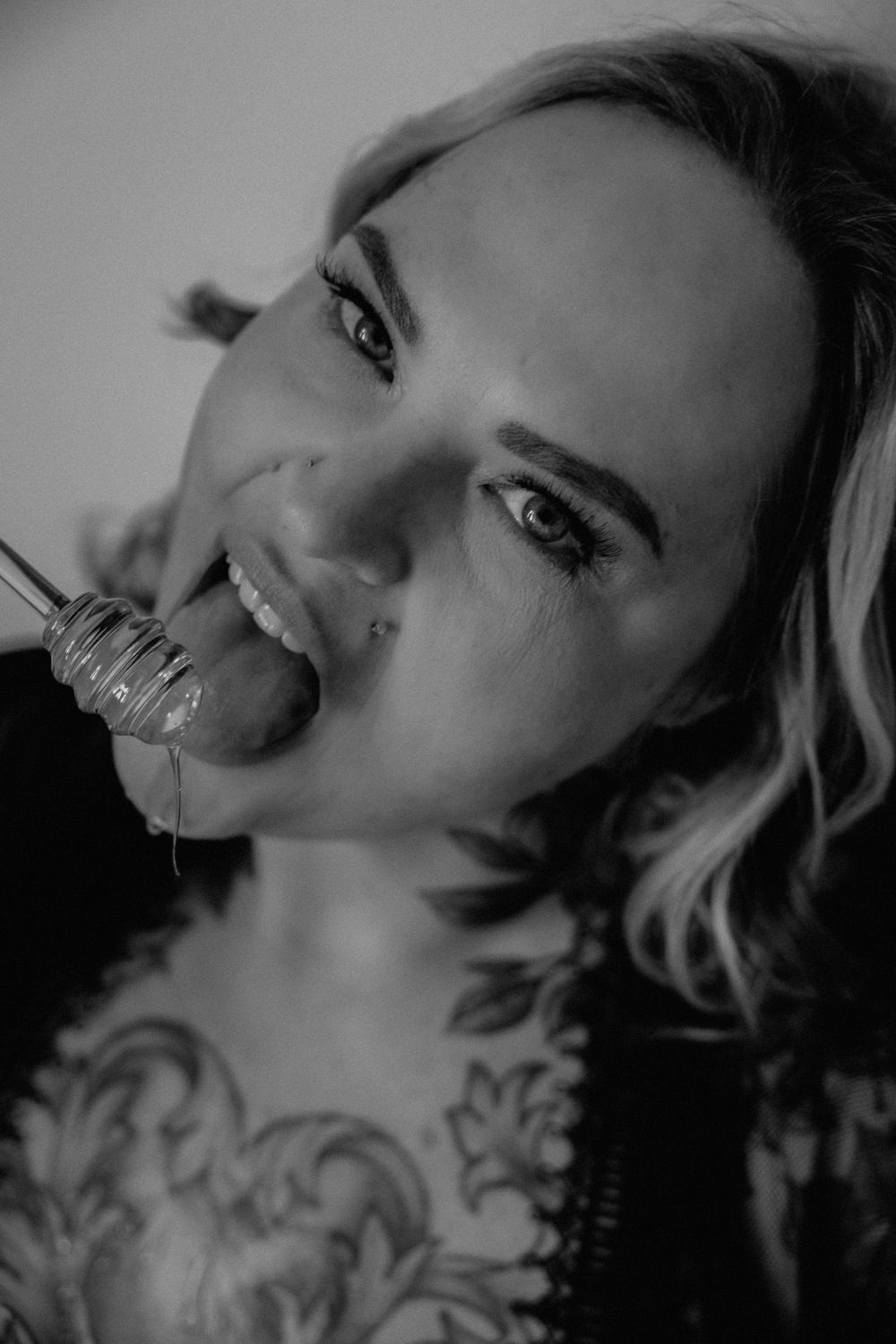 Black and white portrait of tattooed blonde woman licking a spoon with liquid dripping, close-up shot.
