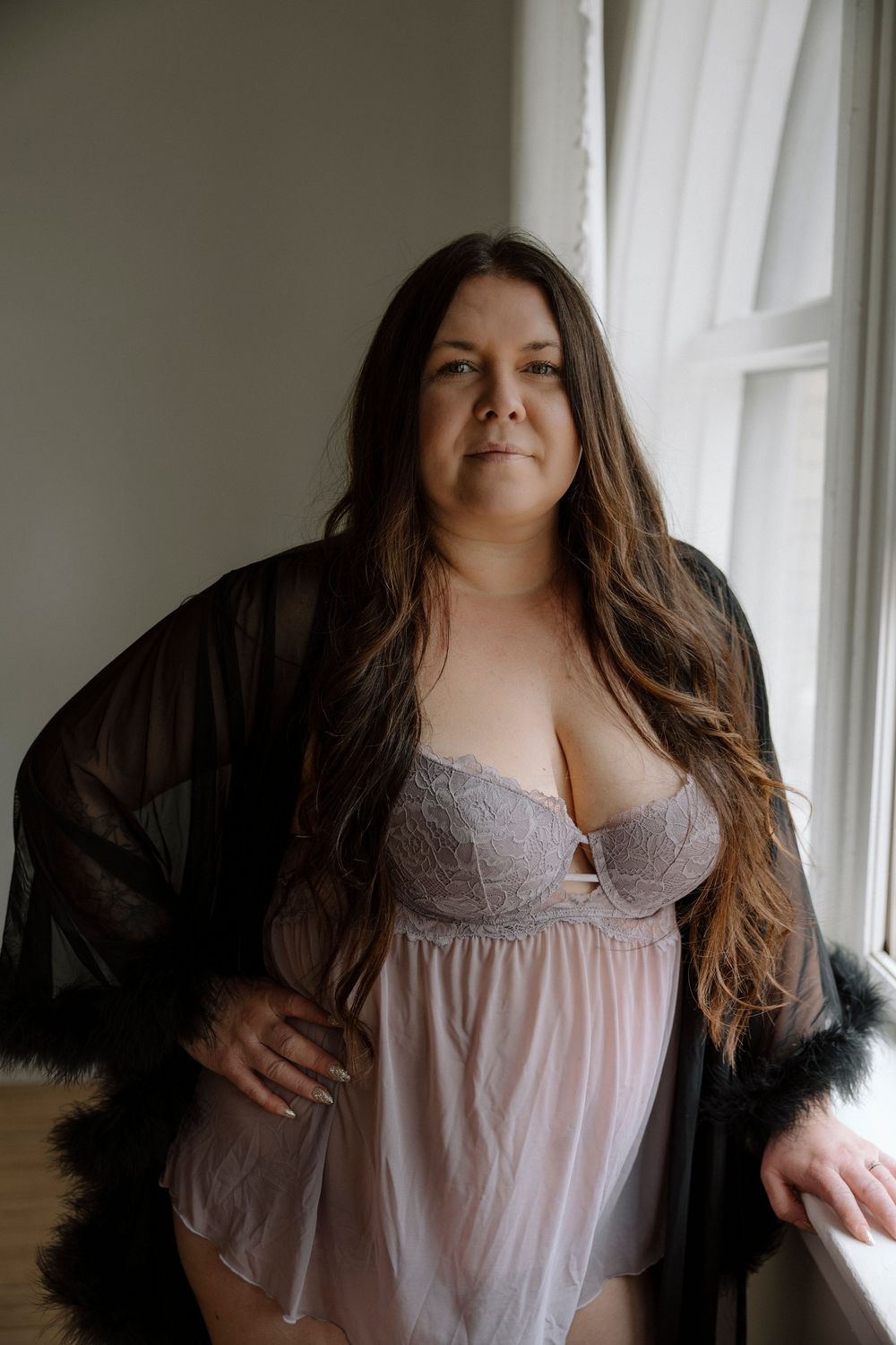 Plus-size woman in pink lace lingerie and sheer black robe poses confidently by a window.