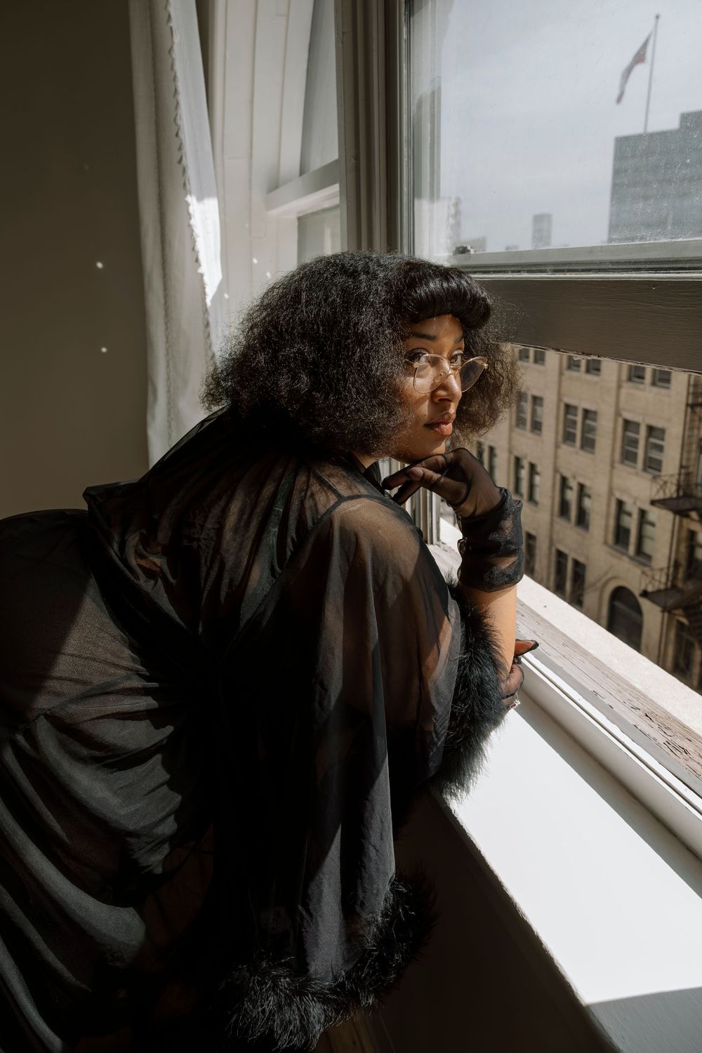 Woman in sheer black robe gazing thoughtfully out a city window, wearing glasses and curly hair.