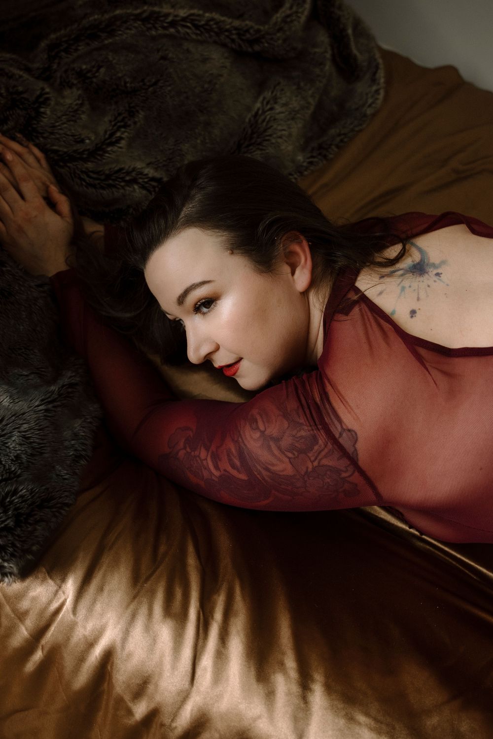 Woman with tattoos in red lace top lying on golden satin sheets with fur blanket in moody boudoir portrait.
