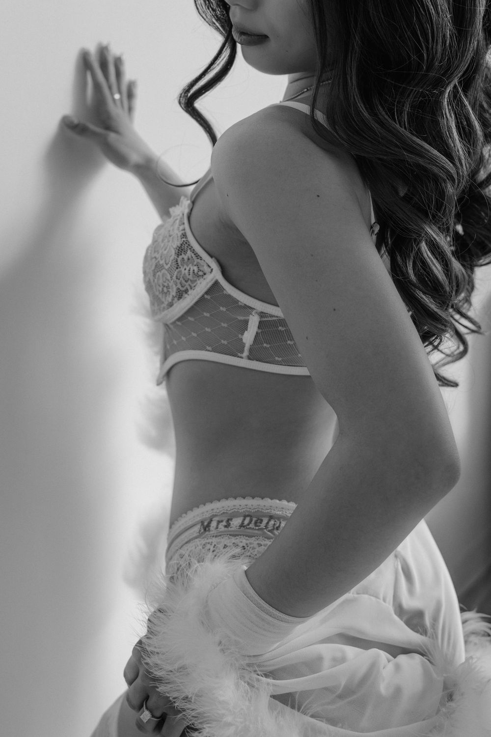 Black and white boudoir photo of a woman in lace lingerie leaning against a white wall with feather accessories.