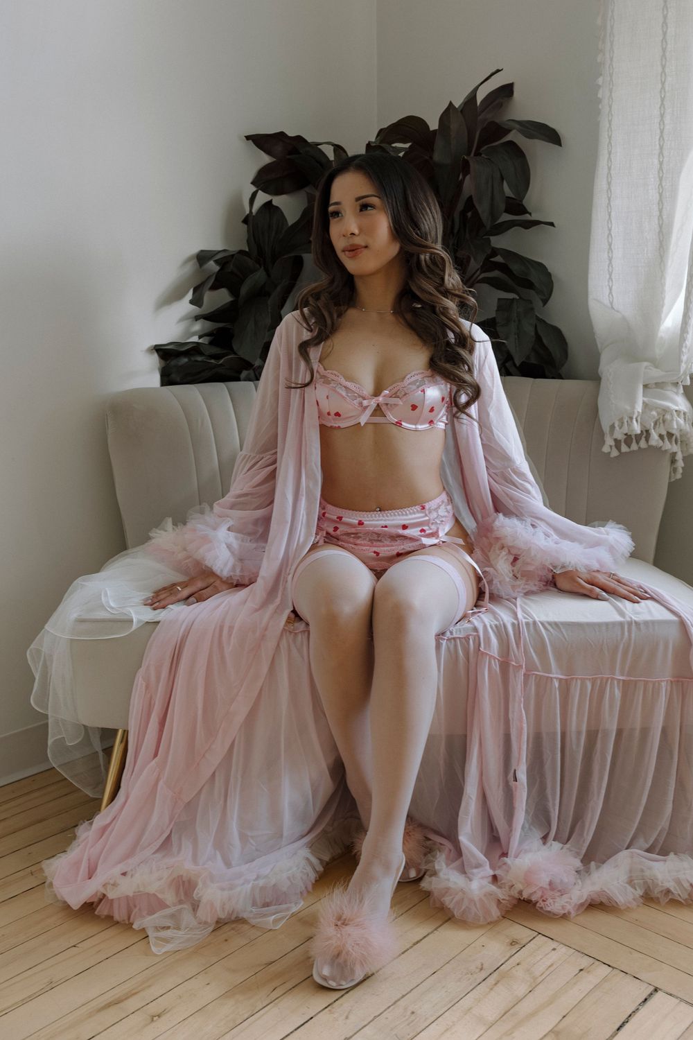 Woman sitting on bed wearing pink lingerie set with sheer robe and fluffy slippers in bright bedroom.