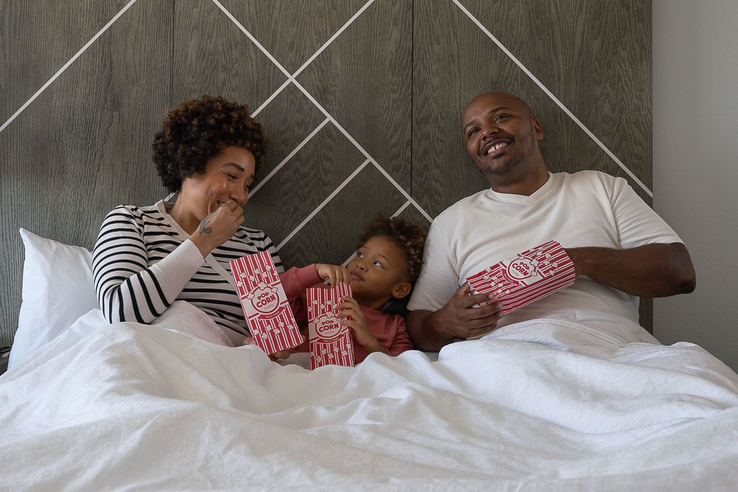 A young family sits in a hotel bed, eating popcorn and laughing.