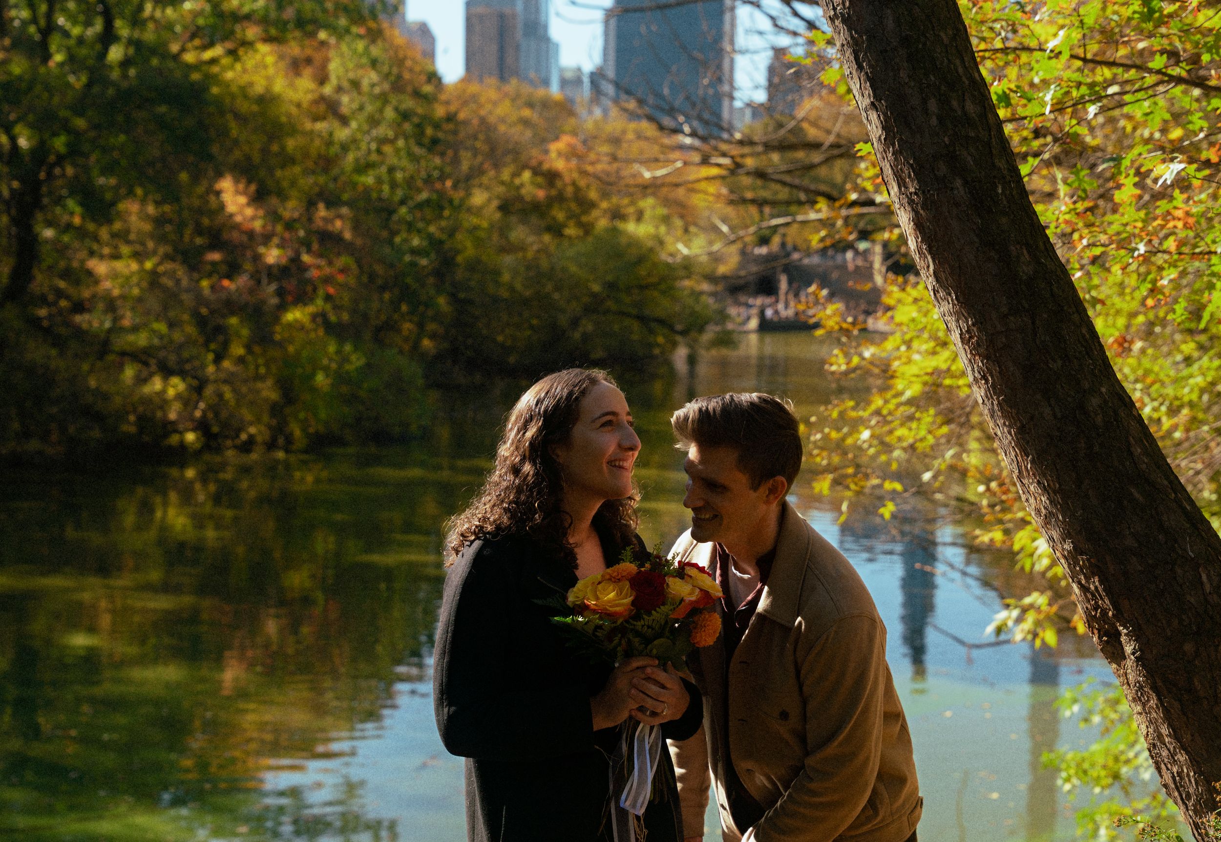 A couple shares a romantic moment on a park path with fall foliage and city skyline in background.