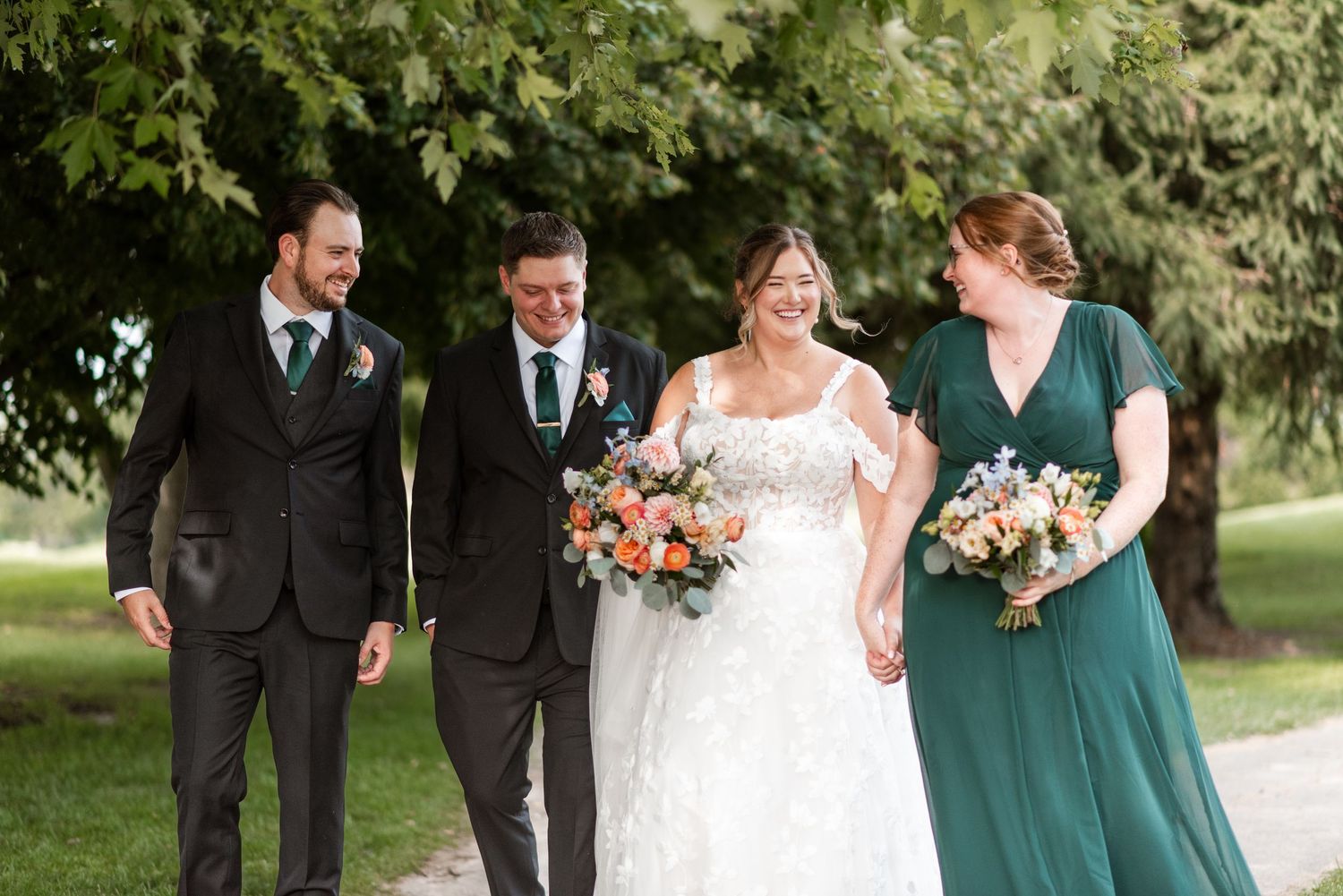 Wedding party in black suits and emerald green dress walk together on garden path holding coral floral bouquets.
