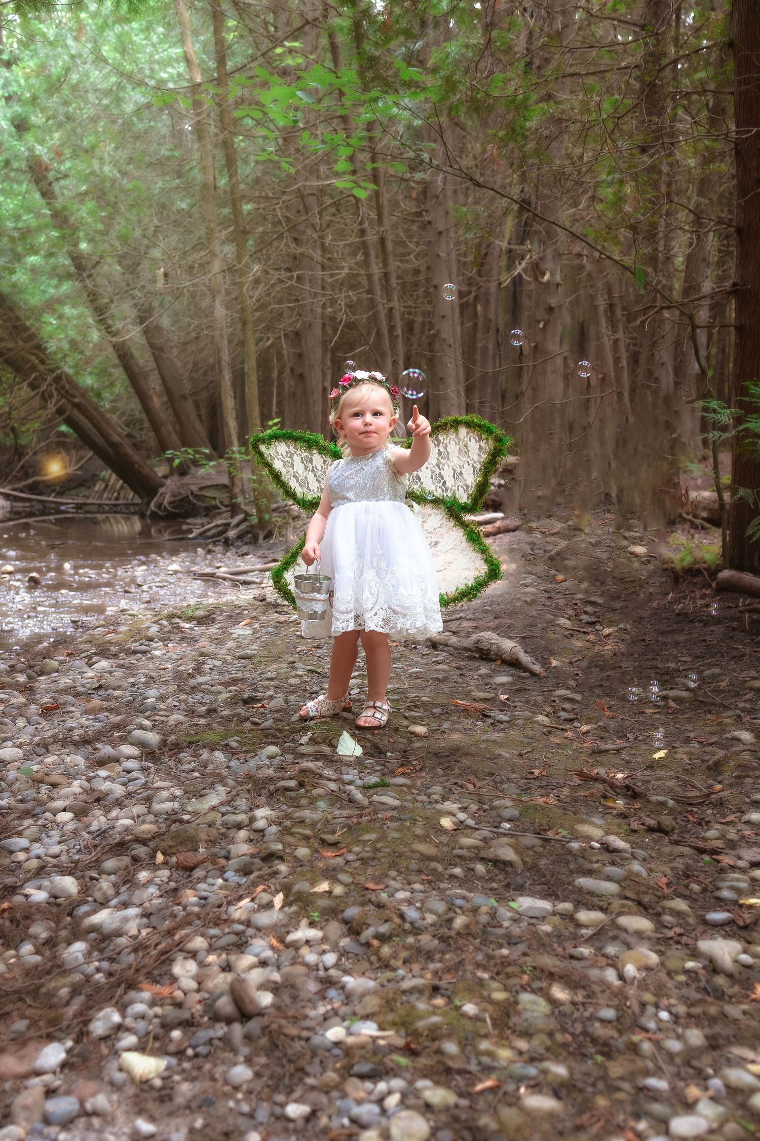 A child wearing white fairy wings poses in a wooded area with scattered leaves on the forest floor.