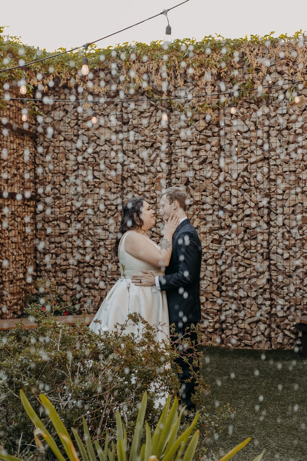 A couple shares a romantic moment in a garden behind a fountain wall at River Garden Weddings and Events.