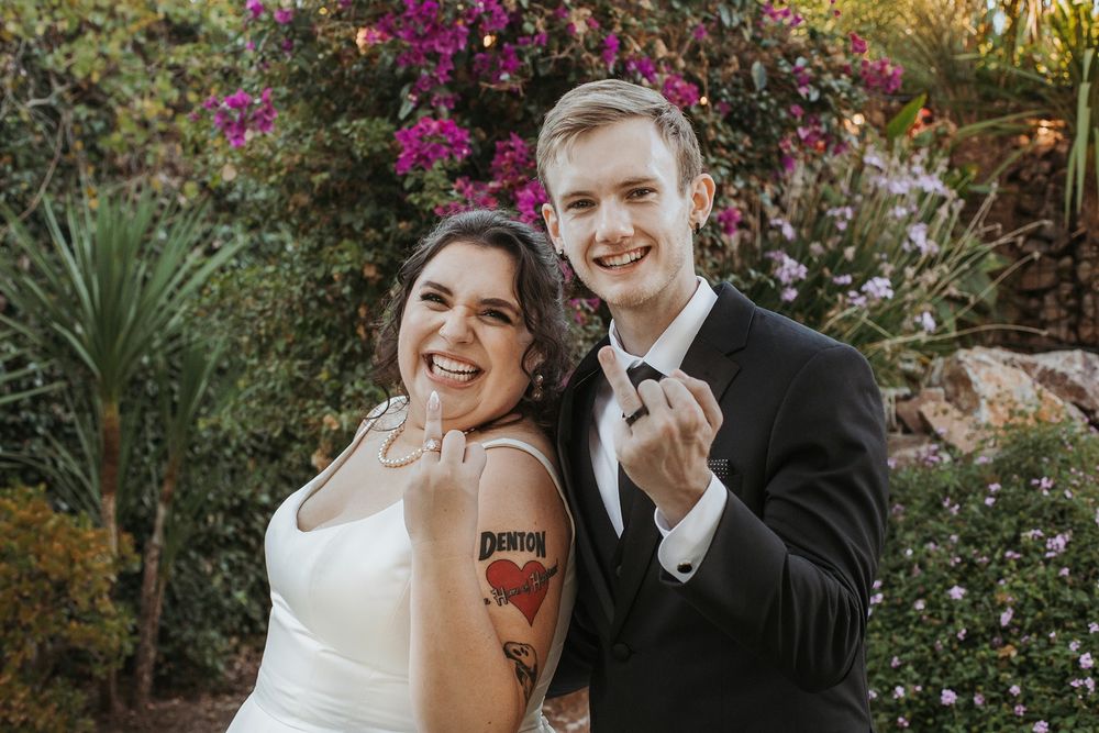 A wedding couple share a fun moment in a lush garden with vibrant pink bougainvillea flowers in bloom at River Garden Weddings and Events.