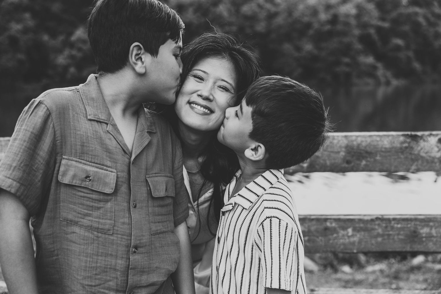 A black and white photo of two boys kissing their laughing mother on the cheeks outdoors.