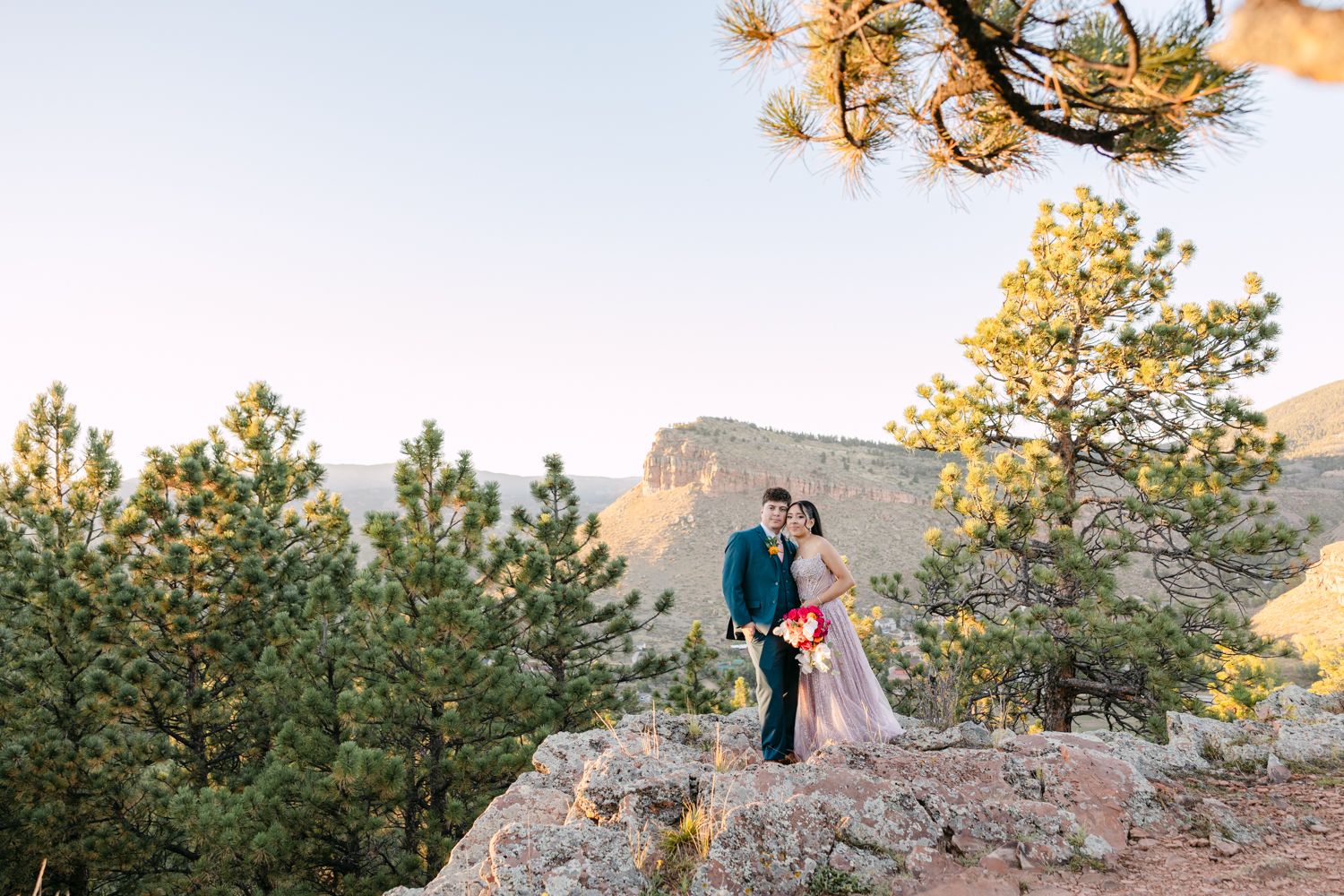 A couple shares romantic moments against a scenic mountain backdrop at sunset while wearing formal attire.