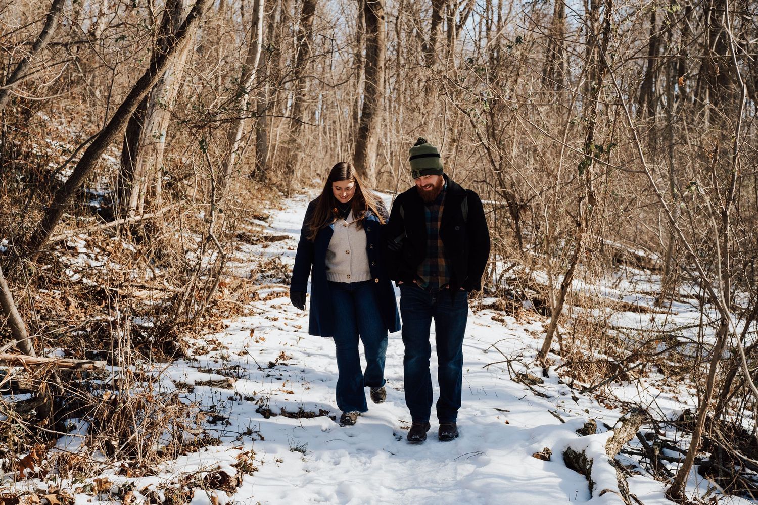 A couple walks together on a snow-covered forest trail during winter for engagement photoshoot in Pequea, PA Lancaster county.