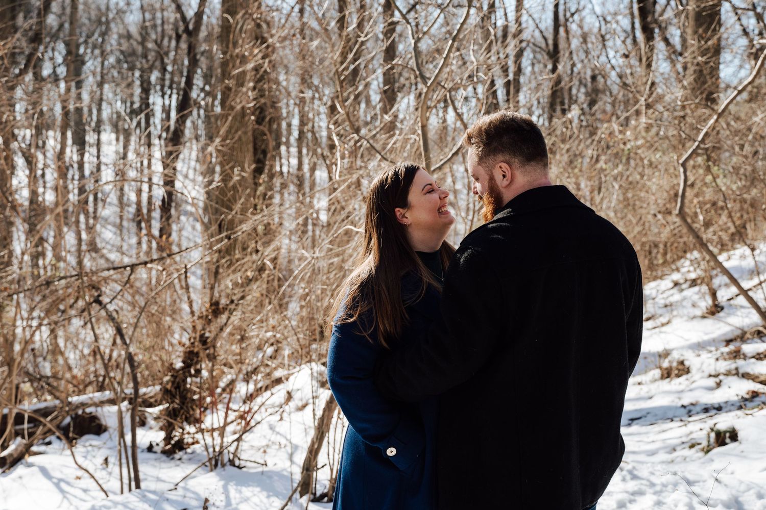A romantic winter scene with a couple embracing among bare trees and snow covered ground for engagement photoshoot in Central PA.
