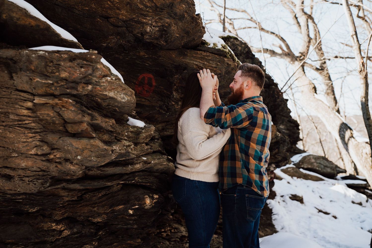A couple embraces while standing in a snowy winter landscape with rocky cliffs in the background.