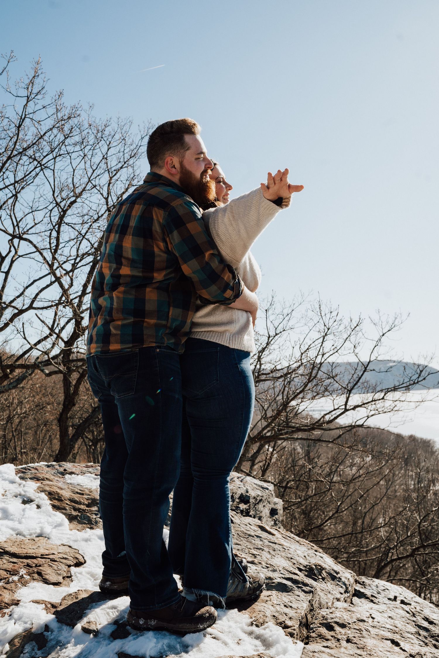 A couple stands together on a snowy mountain overlook, pointing into the distance on a clear winter day.