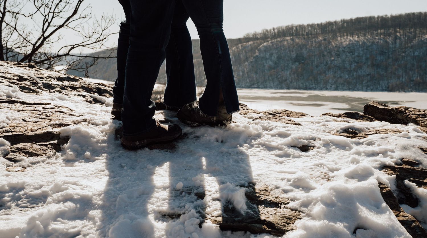 Two pairs of boots stand in the snow at the edge of a frozen lake with a forested shoreline in winter.
