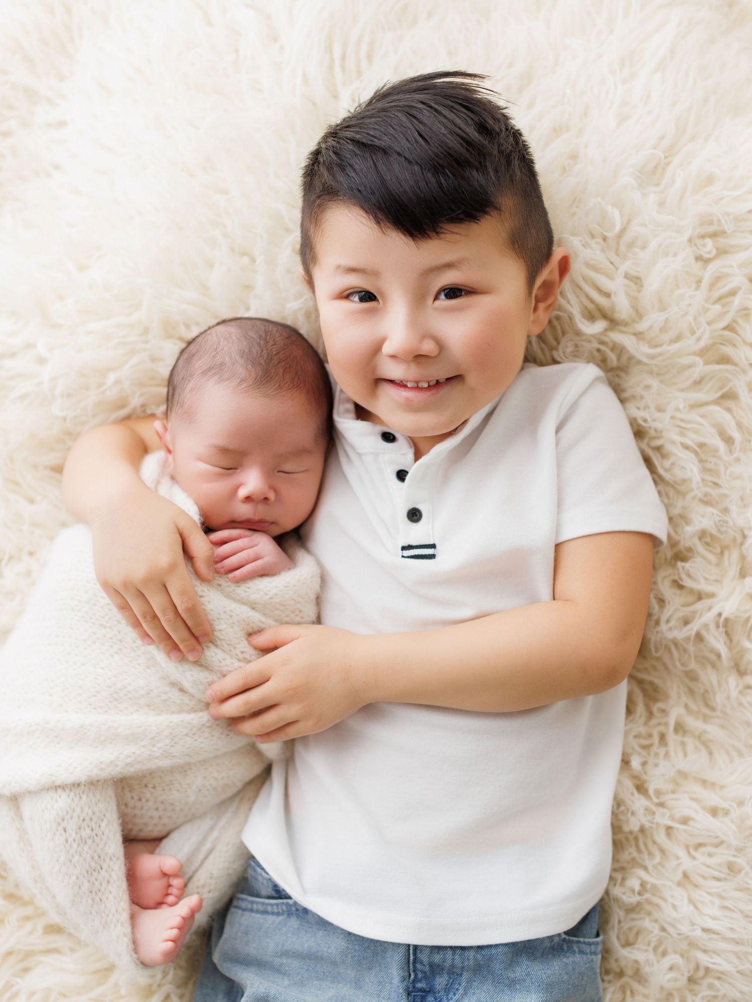Two siblings lying on soft white fur, sharing a tender moment with a sleeping newborn.
