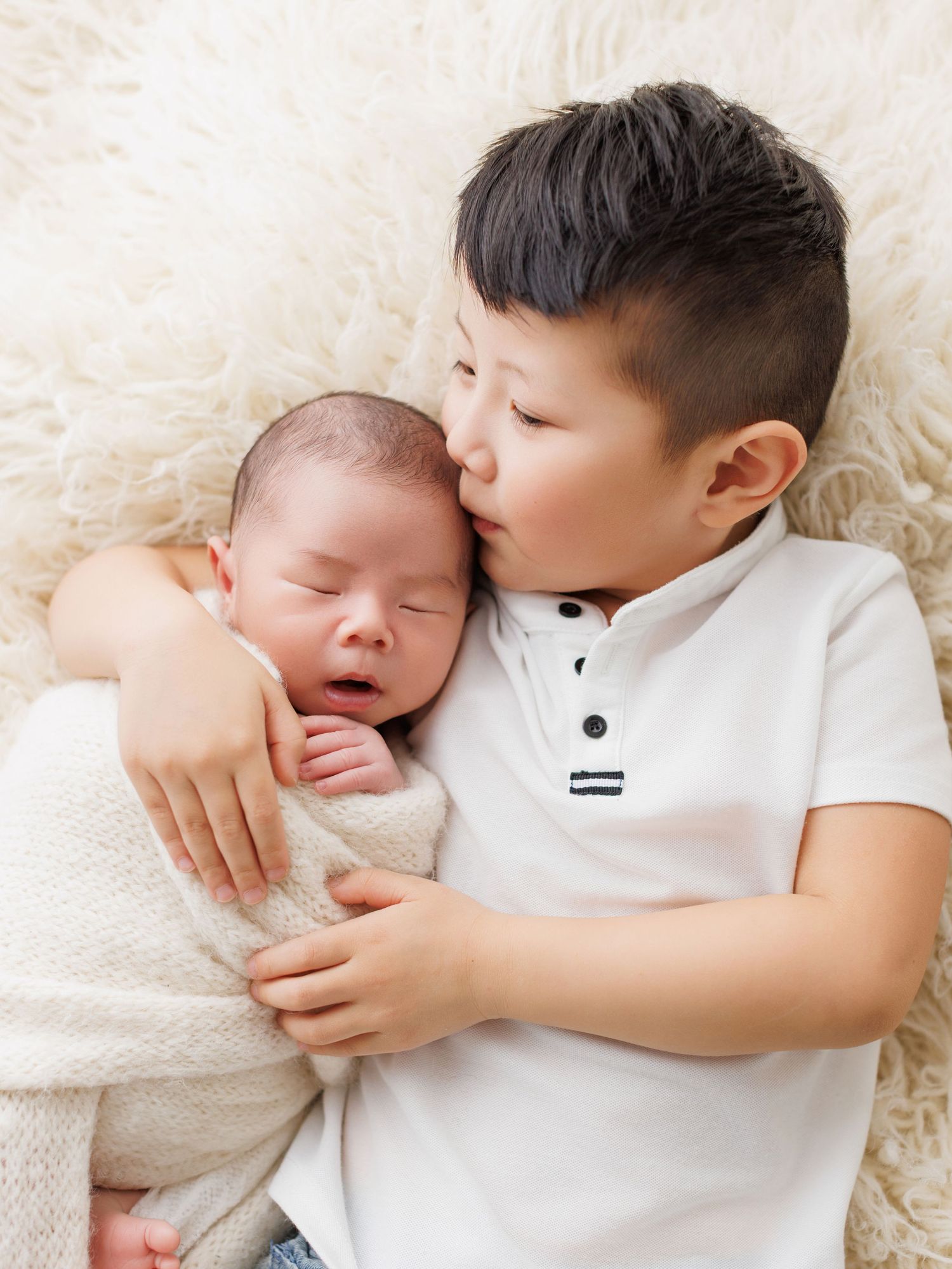A young child in white clothing gently holds and interacts with a sleeping newborn baby on a soft cream blanket.