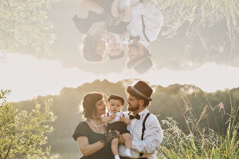 Family portrait with creative reflection effect of parents holding baby in sunny meadow.