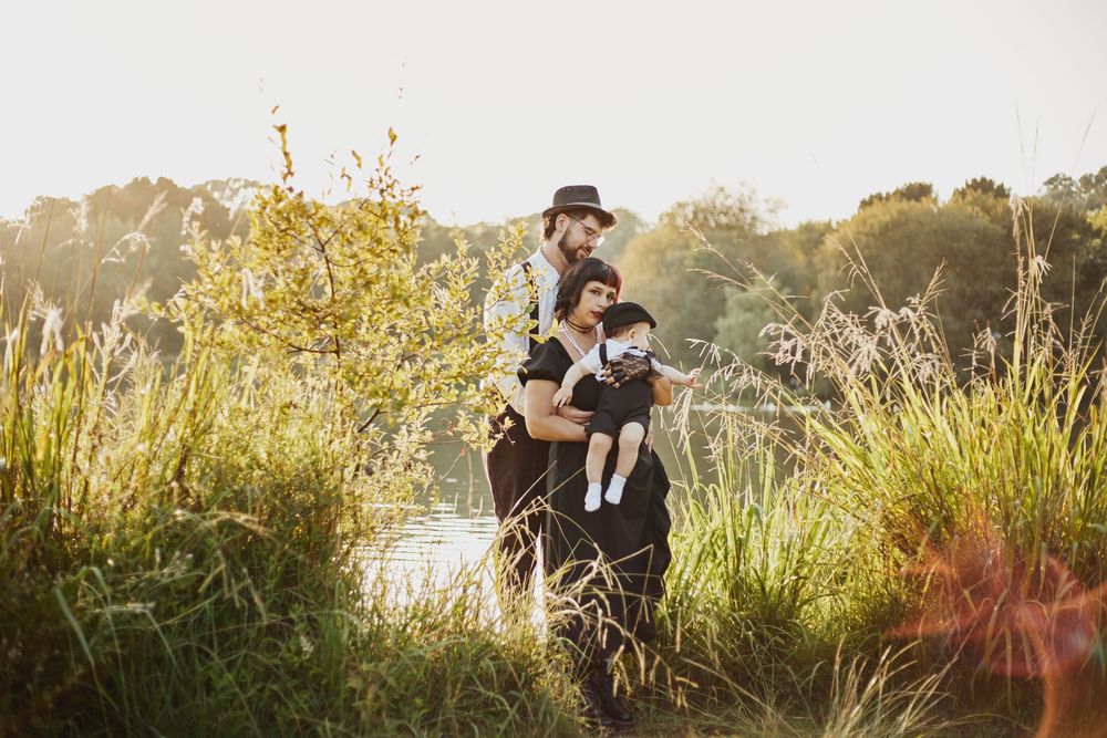 Father holding mother and baby in golden sunset light near river in tall grass.