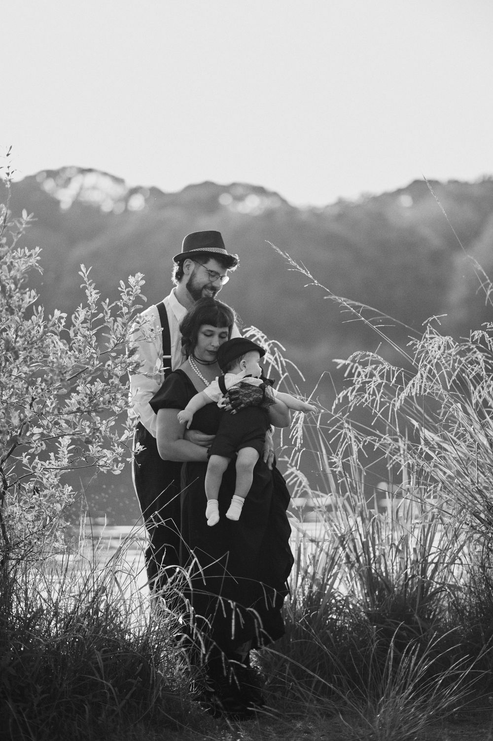 Black and white portrait of bearded father embracing mother holding baby in nature.