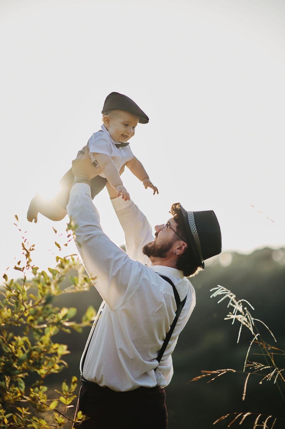 Bearded father lifting laughing baby high in the air at sunset in garden.