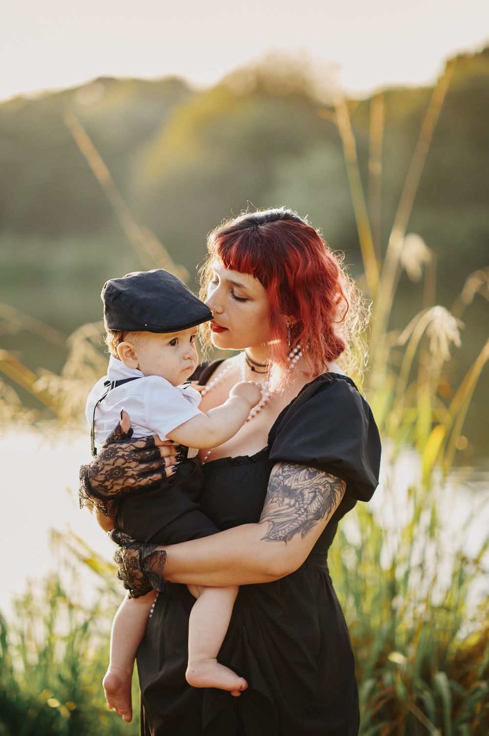 Red-haired tattooed mother in black dress holding baby boy at golden hour by lake.