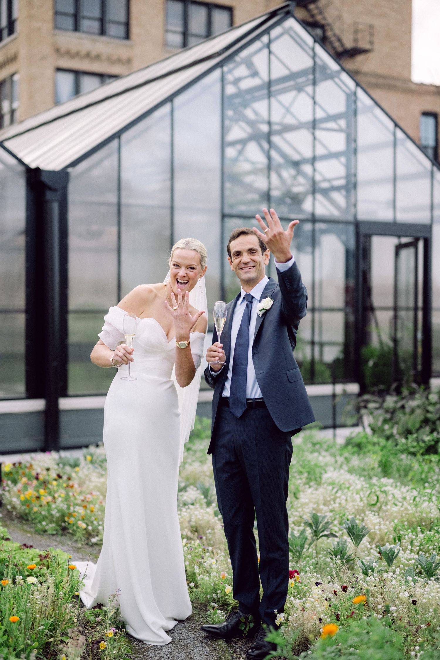 Happy newlywed couple celebrates joyfully in a garden with colorful wildflowers outside a glass building.