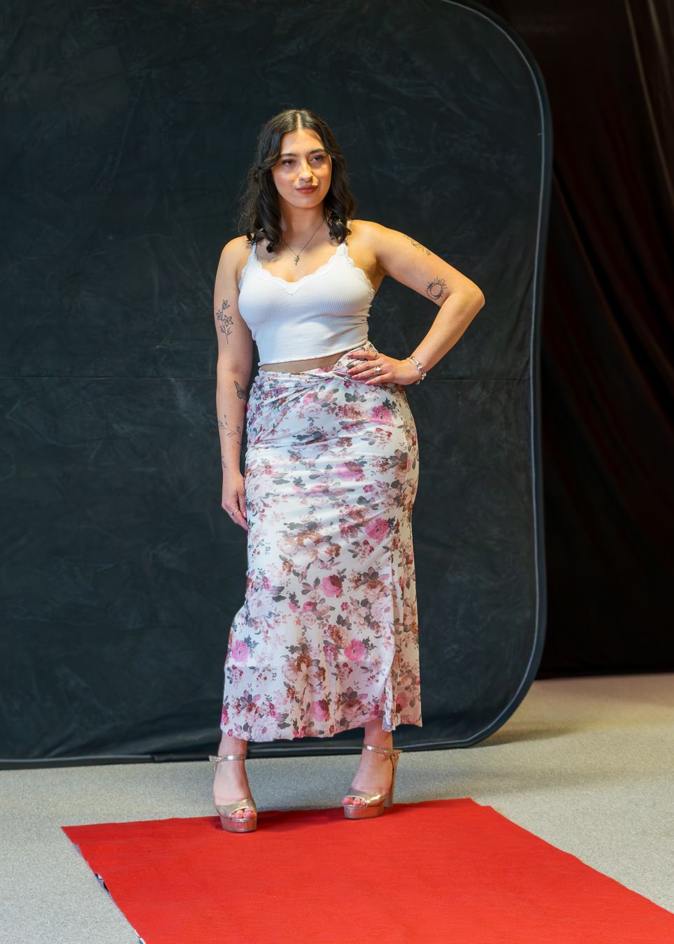 Fashion model in white top and floral skirt poses on red carpet against dark backdrop during photoshoot.