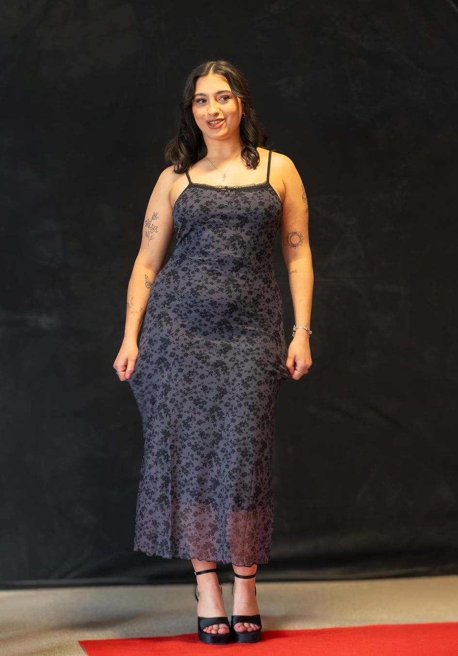 A person wearing a navy floral dress stands on a dark stage with red carpet, showing different angles of the outfit.