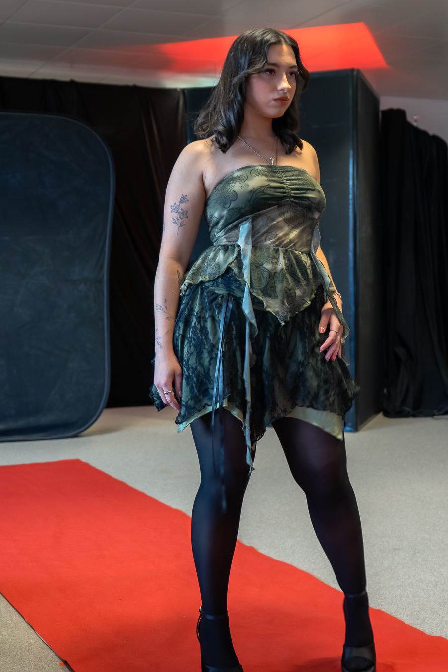 Model wearing dark green velvet dress strikes poses on red carpet runway against black background.