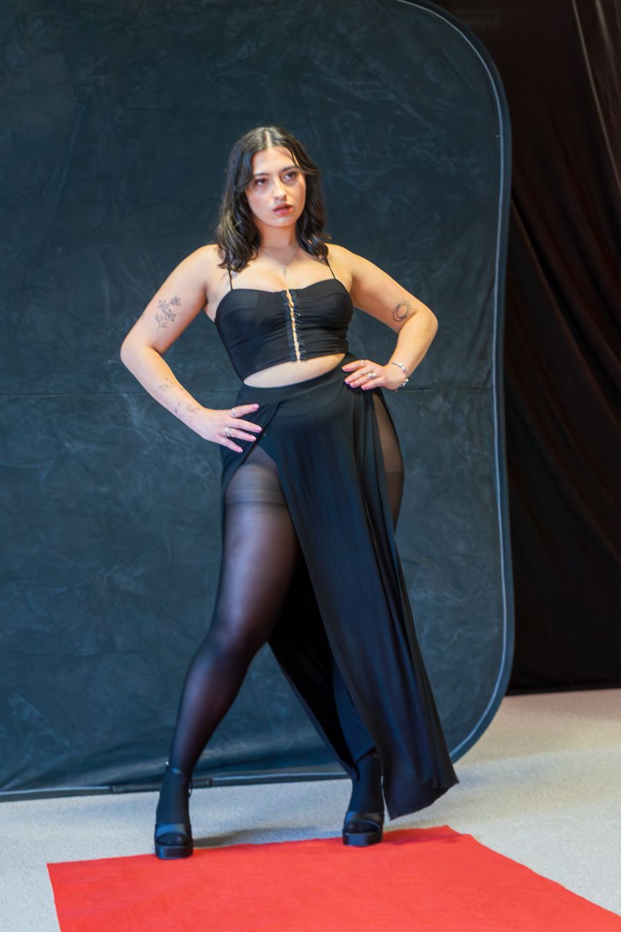 Model in black dress and tights poses dramatically on red carpet during fashion photography session.