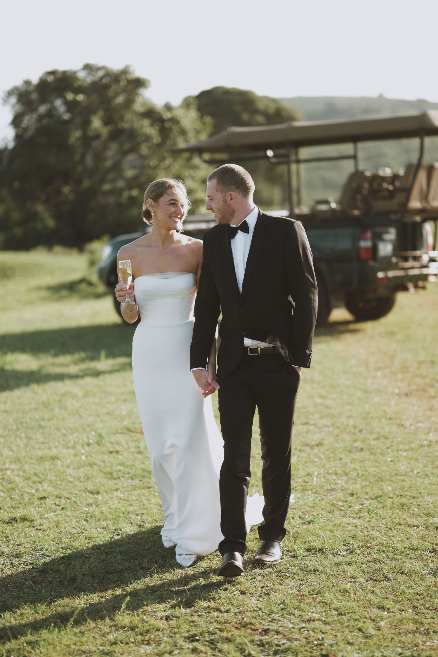 Bride in white strapless gown and groom in black tuxedo walking hand in hand at golden hour near safari vehicle.