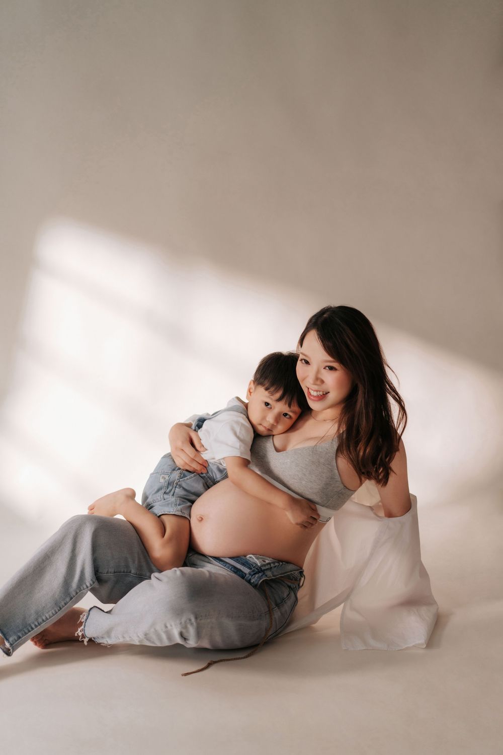 Artistic maternity photos in soft lighting show a mother and child reclining on white fabric in gentle poses.
