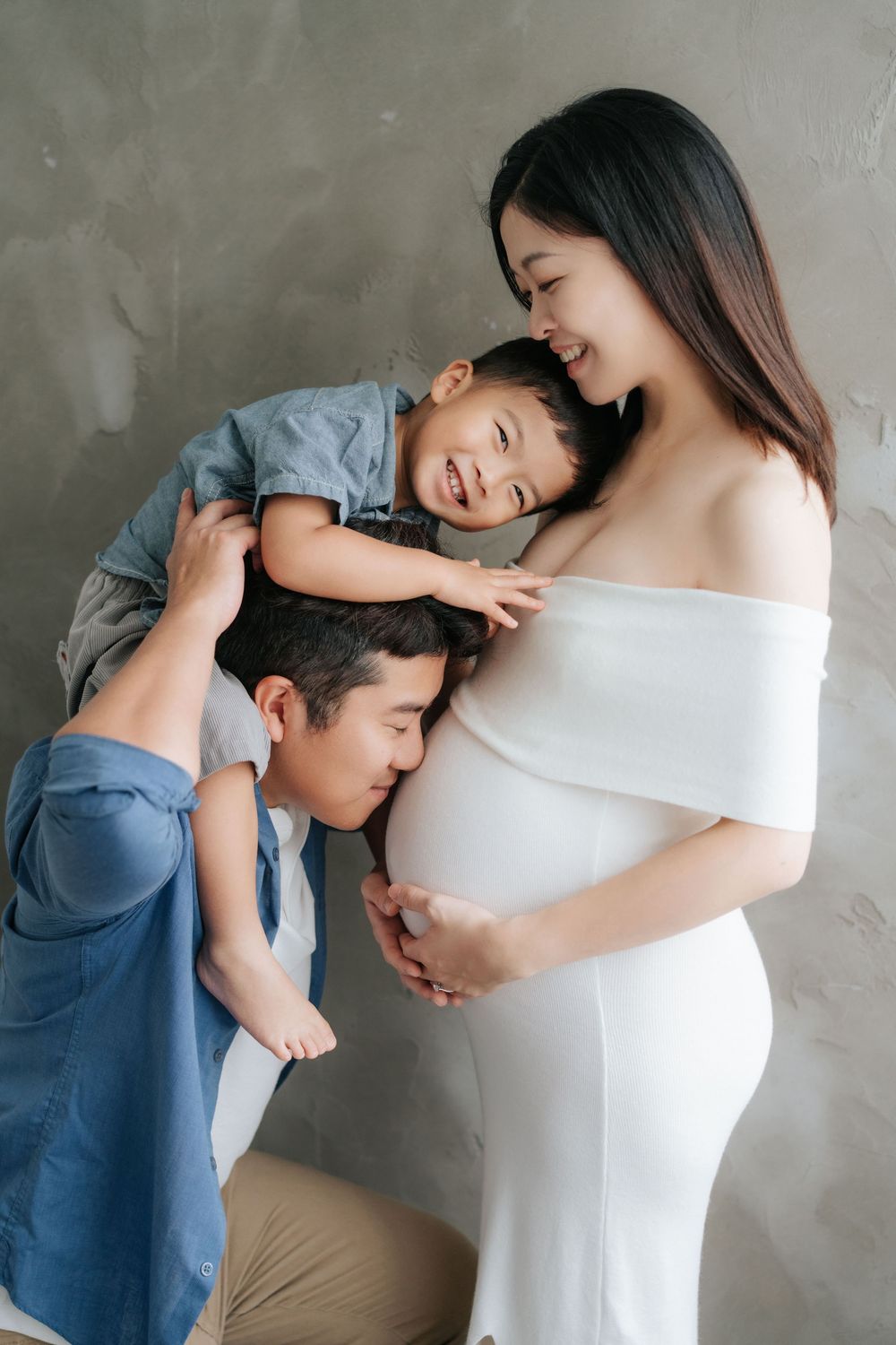 A tender family moment with an expectant mother in a white dress while a child playfully rests on shoulders against gray wall.